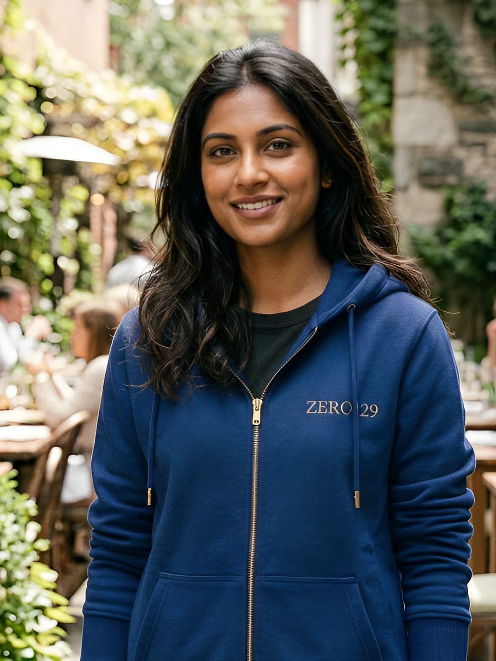 Model in the cobalt Zero129 zip hoodie with gold embroidery, in a garden courtyard